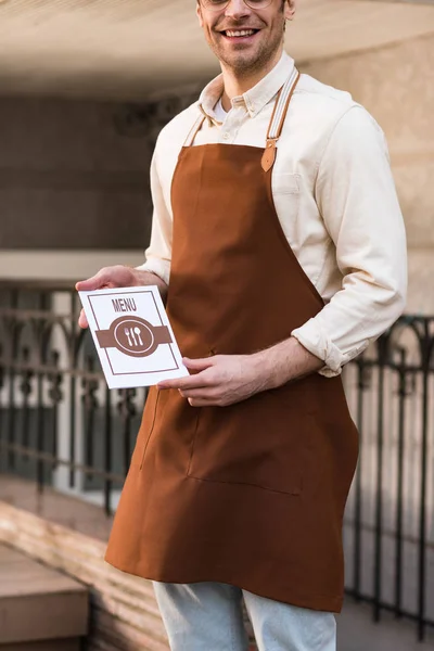 Vue recadrée du barista souriant dans le tablier tenant le menu dans la rue — Photo de stock