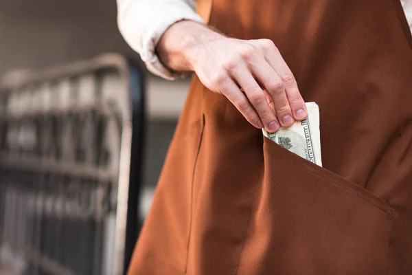 Частичный вид баристы в коричневом фартуке кладет деньги в карман — стоковое фото