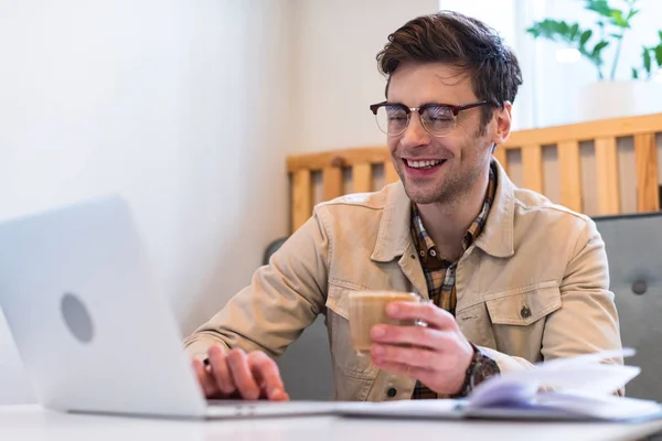 Улыбающийся фрилансер в очках, держа чашку кофе и печатая на клавиатуре ноутбука — стоковое фото