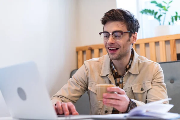 Freelancer sorridente em óculos segurando xícara de café e digitando no teclado do laptop — Fotografia de Stock