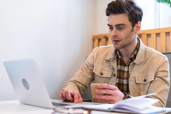 Freelancer concentrado sosteniendo taza de café y usando laptop - foto de stock