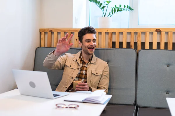 Excited freelancer with laptop holding cup of coffee and waving hand in cafe — Stock Photo