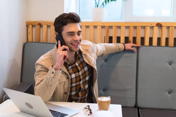 Cheerful freelancer in jacket talking on smartphone in cafe — Stock Photo