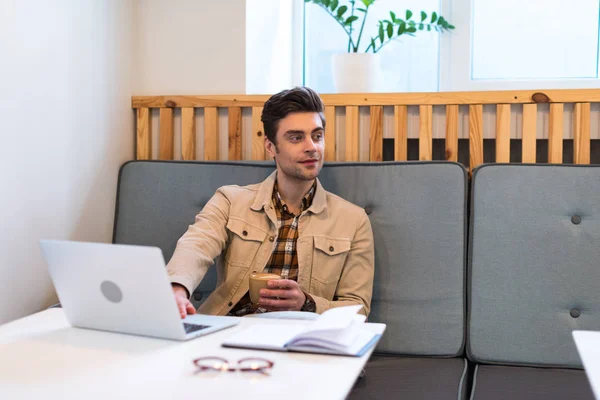 Freelancer with laptop holding cup of coffee in cafe — Stock Photo