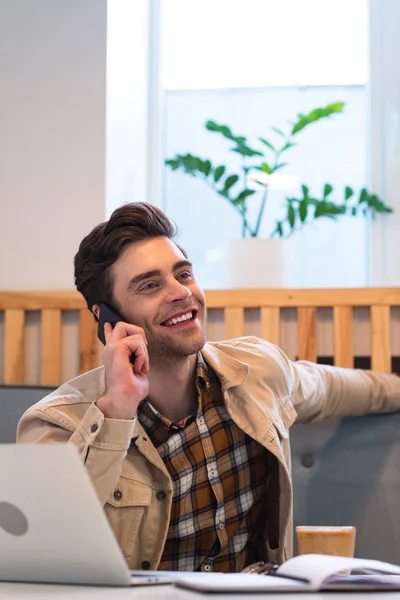 Cheerful freelancer in jacket talking on smartphone in cafe — Stock Photo