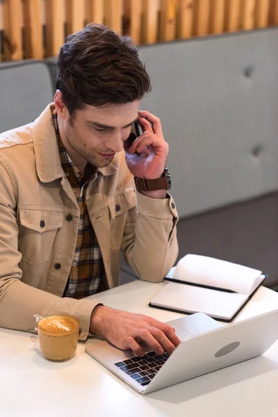 Smiling freelancer in jacket talking on smartphone in cafe — Stock Photo