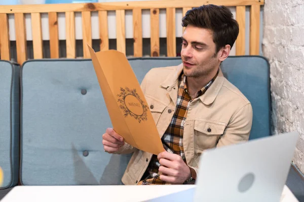 Freelancer in jacket with laptop reading menu in cafe — Stock Photo