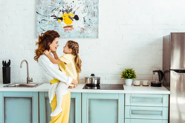 Vista lateral de madre feliz sosteniendo a hija excitada en la cocina - foto de stock