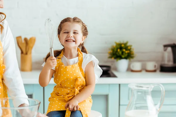 Animado filha no bolinhas ponto amarelo avental segurando balão batedor ao lado da mãe na cozinha — Fotografia de Stock