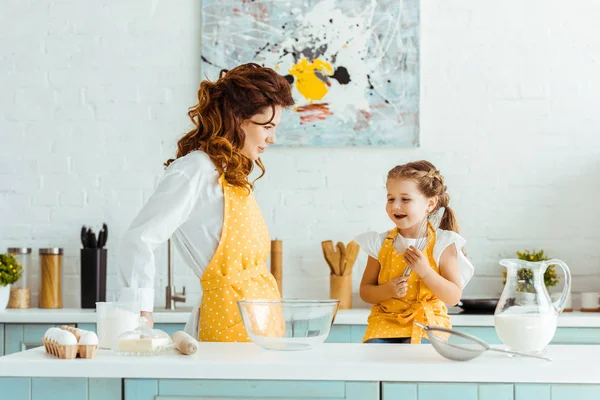 Madre en delantal amarillo lunar mirando emocionada hija en la cocina - foto de stock