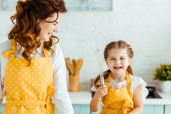 Smiling mother looking at happy daughter with balloon whisk in kitchen — Stock Photo