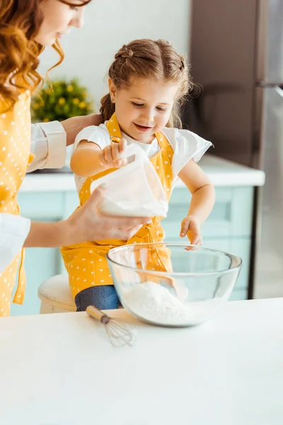 Mãe em bolinhas ponto avental amarelo ajudando a filha sorrindo adicionando farinha para tigela — Fotografia de Stock