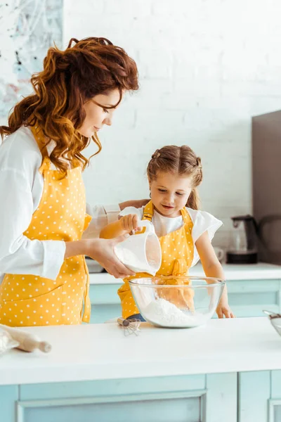 Madre in pois grembiule giallo aiutare figlia attenta l'aggiunta di farina a ciotola — Foto stock