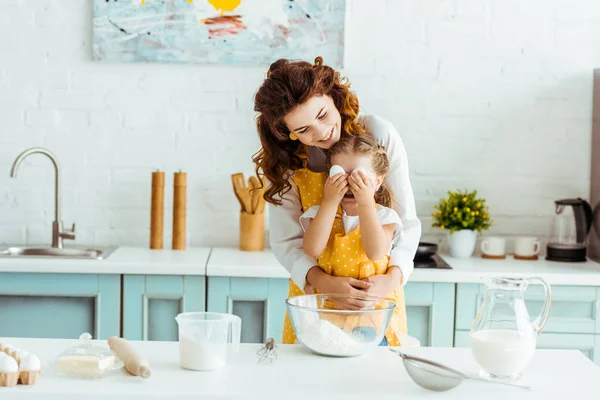 Mère heureuse embrassant fille tandis que l'enfant tenant des œufs devant les yeux dans la cuisine — Photo de stock