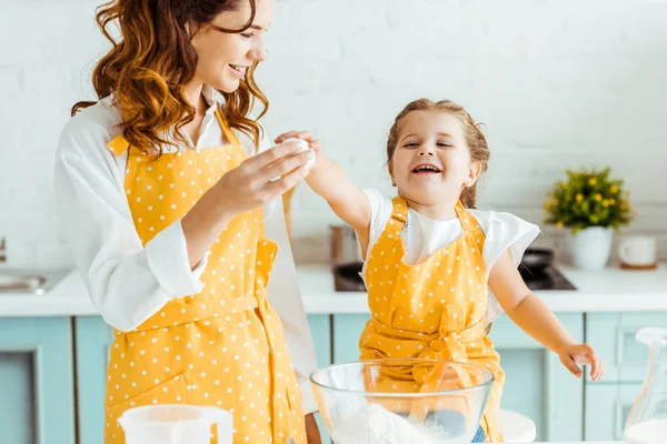 Excited emotional daughter holding egg with mother in yellow polka dot apron in kitchen — Stock Photo