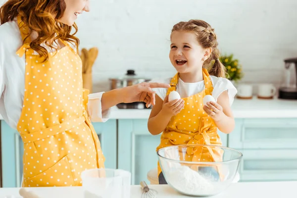 Eccitata figlia emotiva che tiene le uova mentre la madre in grembiule a pois giallo punta con il dito in cucina — Foto stock