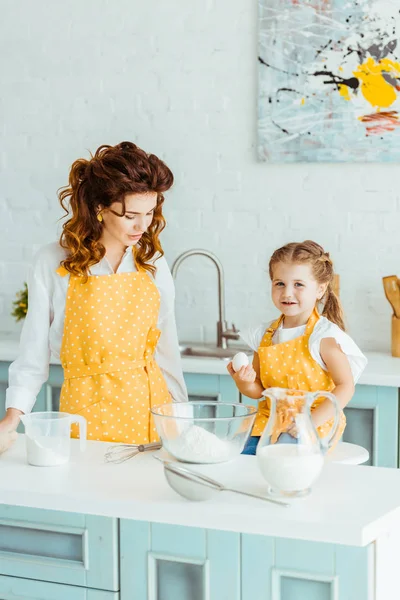 Cute kid in yellow polka dot apron holding egg near mother in kitchen — Stock Photo