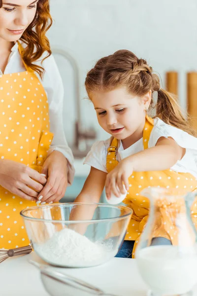 Filha bonito adicionando ovo à farinha na tigela enquanto cozinha com a mãe na cozinha — Fotografia de Stock