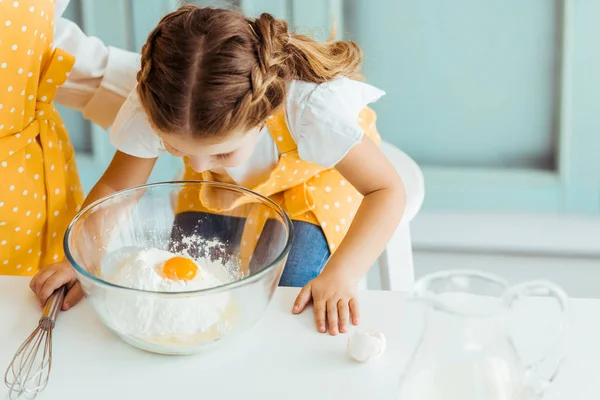 Cute kid in apron looking at smashed egg on flour in bowl on table — Stock Photo