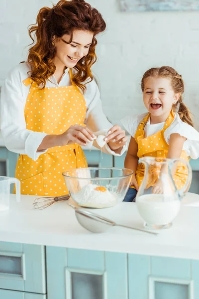 Glückliche Mutter und Tochter fügen zertrümmertes Ei zu Mehl in Schüssel hinzu, während sie gemeinsam in der Küche kochen — Stockfoto