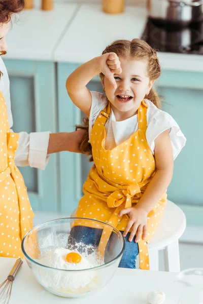 Mother touching daughter in apron near bowl with flour and smashed egg while excited child showing thumb down — Stock Photo