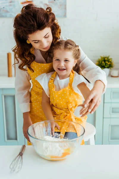 Mutter und aufgeregte Tochter in gelben Tupfen-Schürzen neben Schüssel mit Mehl und Eiern auf dem Tisch — Stockfoto