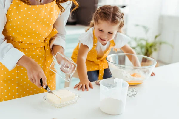 Mulher em amarelo polka dot avental de corte de manteiga perto feliz filha na cozinha — Fotografia de Stock