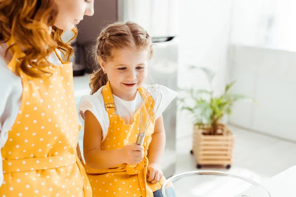 Mother looking at happy daughter in polka dot apron with balloon whisk — Stock Photo