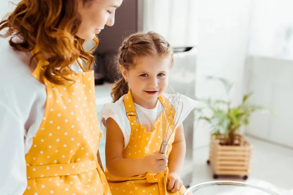 Mère regardant fille mignonne à pois tablier avec ballon fouet — Photo de stock