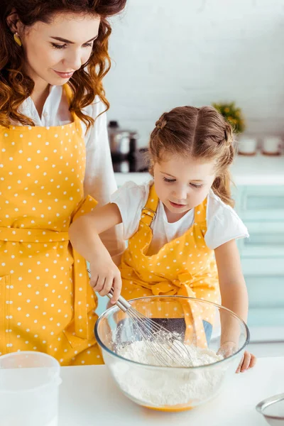 Mutter schaut süße Tochter in gepunkteter Schürze an, die Mehl und Eier mit Luftballonbesen vermischt — Stockfoto