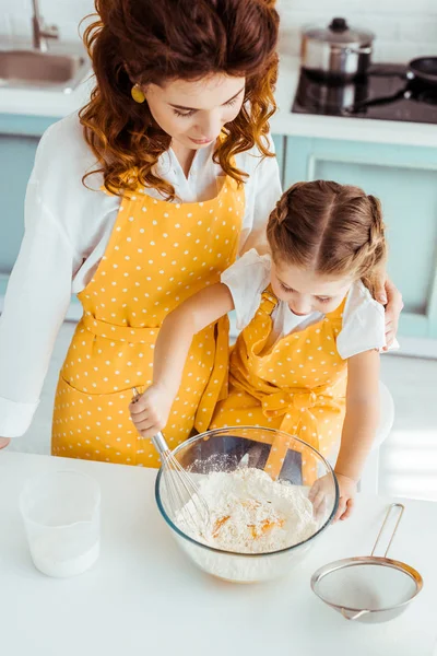 Vista aerea della madre guardando carina figlia in grembiule a pois mescolando farina e uova con frusta palloncino — Foto stock