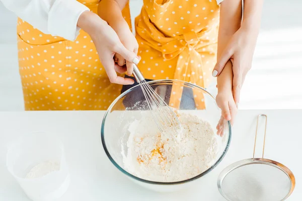 Vista recortada de la madre y la hija en delantales de lunares mezclando harina y huevos con batidor de globo en un tazón - foto de stock