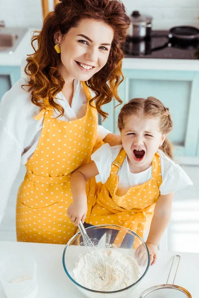 Overhead view of mother and excited daughter in polka dot aprons mixing flour and eggs with balloon whisk in bowl — Stock Photo