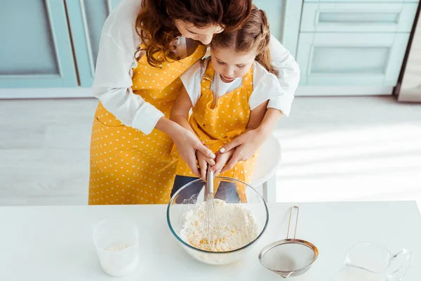 Vista aérea de la madre y la hija en delantales mezclando harina y huevos con batidor de globo en un tazón - foto de stock