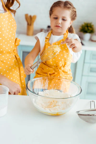 Selective focus of daughter in yellow polka dot apron looking at balloon whisk with dough — Stock Photo
