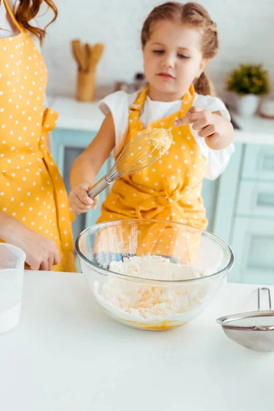 Selektiver Fokus der Tochter in gelber Tupfenschürze mit Blick auf Ballonbesen mit Teig — Stockfoto