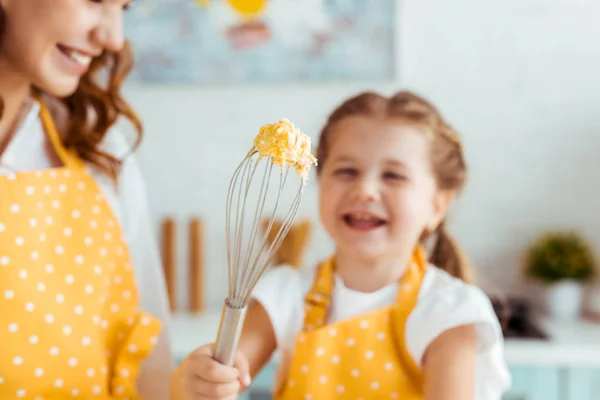 Selective focus of smiling mother and laughing daughter holding balloon whisk with raw dough — Stock Photo