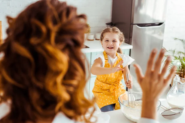 Enfoque selectivo de niño emocionado en delantal de masa para cocinar en la cocina y mirando a la madre saludando la mano - foto de stock