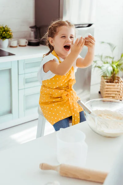 Excited kid in yellow polka dot apron looking at eggs while cooking dough in kitchen — Stock Photo