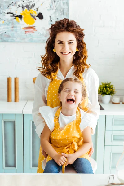 Feliz madre y riendo hija en lunares delantales amarillos en la cocina - foto de stock