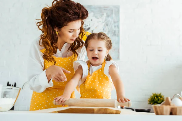 Daughter in polka dot apron rolling out dough on baking parchment paper while mother pointing with finger on it — Stock Photo