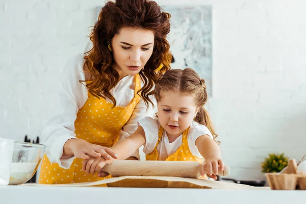 Madre joven ayudando a su hija a desplegar masa en papel de pergamino para hornear - foto de stock