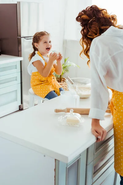 Excitada hija en delantal de lunares mirando a la madre en la cocina - foto de stock