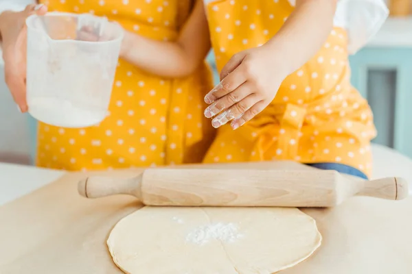 Cropped view of mother and daughter in yellow polka dot aprons holding measuring jug with flour near dough and wooden rolling pin on baking parchment paper — Stock Photo