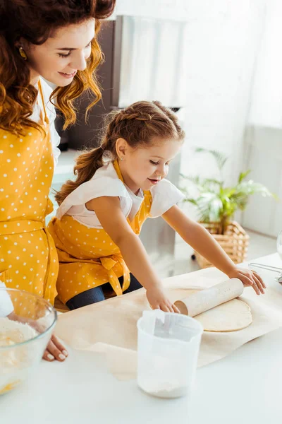 Glückliche Tochter in gelber Tupfen-Schürze rollt Teig auf Tisch neben Mutter aus — Stockfoto