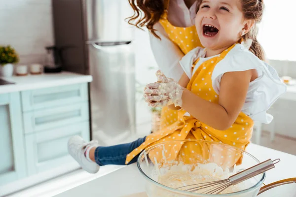 Excited child in polka dot apron with dough on hands laughing while sitting next to mother on kitchen table — Stock Photo