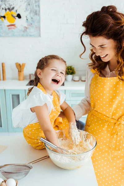 Lächelnde Mutter schaut lachende glückliche Tochter mit Händen in Schüssel mit Teig an — Stockfoto