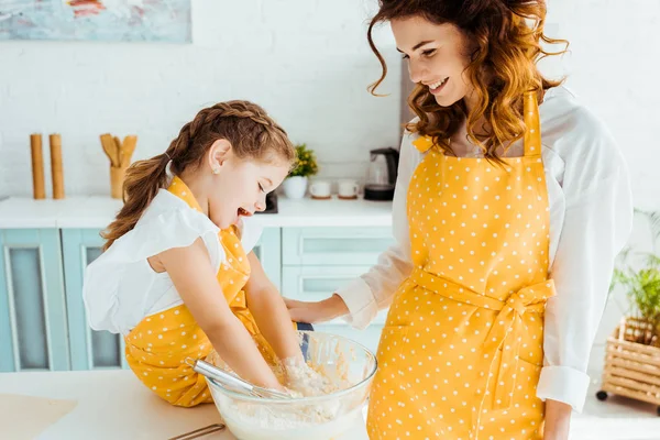 Mère souriante regardant fille heureuse mettre les mains dans un bol avec de la pâte — Photo de stock