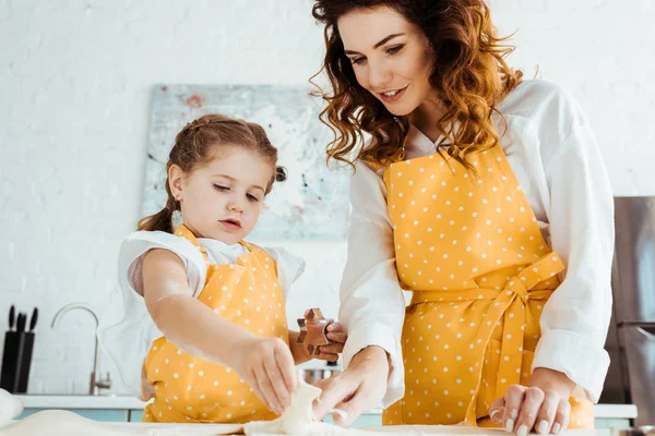 Усміхнена мати в жовтому точковому фартуху, дивлячись на дочку, що тримає тісто на кухні — стокове фото