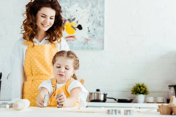 Mère heureuse en tablier à pois jaunes regardant fille tenant des moules à pâte dans la cuisine — Photo de stock